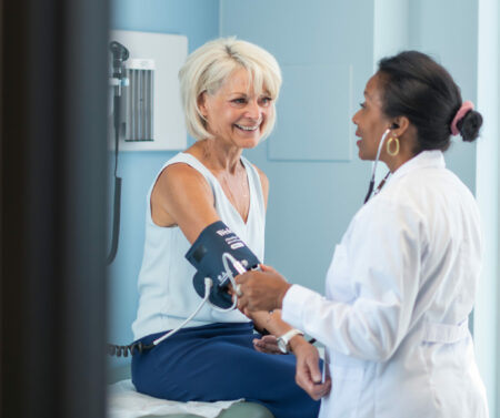 A mature adult woman is at a routine medical appointment. Her healthcare provider is a black woman. The patient is sitting on an examination table in a clinic. She is smiling at the doctor. The doctor is checking the patient's blood pressure.